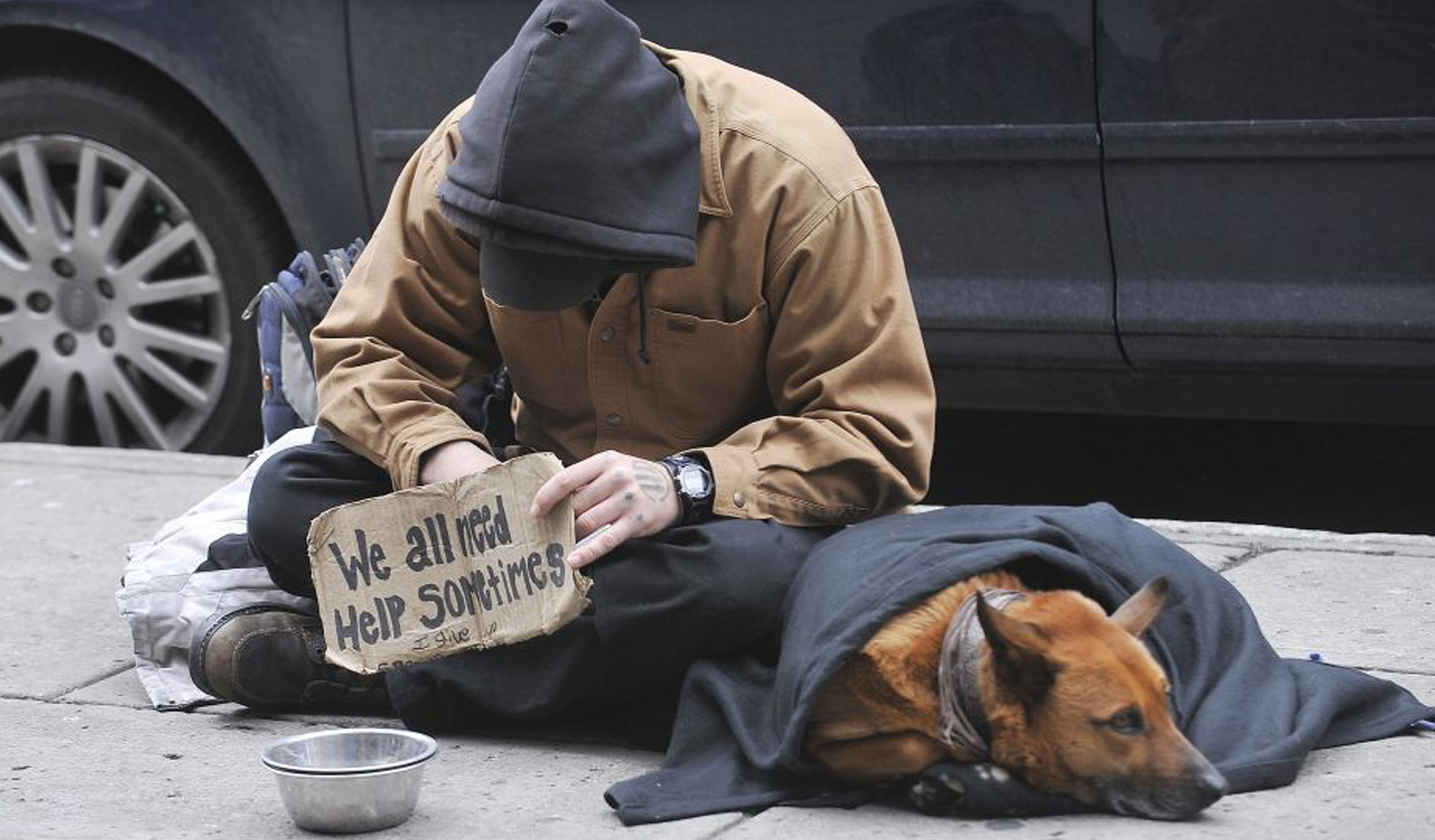 man sitting on the street, leaned over, next to dog, holding a sign